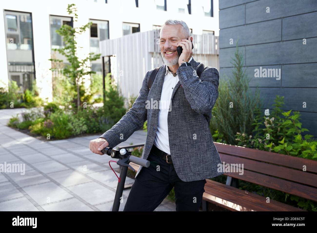 Fröhliche mittleren Alters Geschäftsmann in stilvollen Anzug lächelnd, während am Telefon zu sprechen, stehen mit Elektroroller im Freien Stockfoto