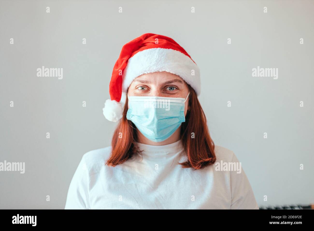 Frau in santa Hut und schützende medizinische Maske auf grauem Wandhintergrund. Neujahr in Quarantäne während der Coronavirus-Pandemie Stockfoto