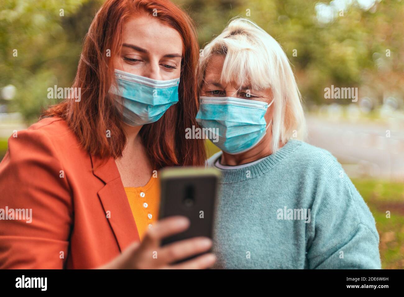 Coronavirus, Covid 19. Mädchen und Erwachsene Frau in Maske verwenden Smartphone-App auf der Stadt Straße. Menschen, die Videos auf der Straße im Park ansehen Stockfoto