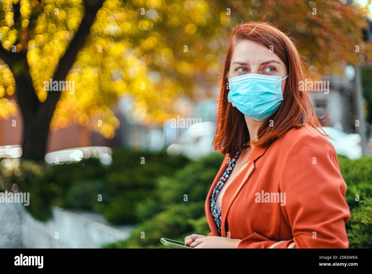 Coronavirus, covid 19 Konzept. Junges Mädchen in medizinischer Maske und Anzug hält ein Smartphone in den Händen und schaut zur Seite auf der Straße Stockfoto