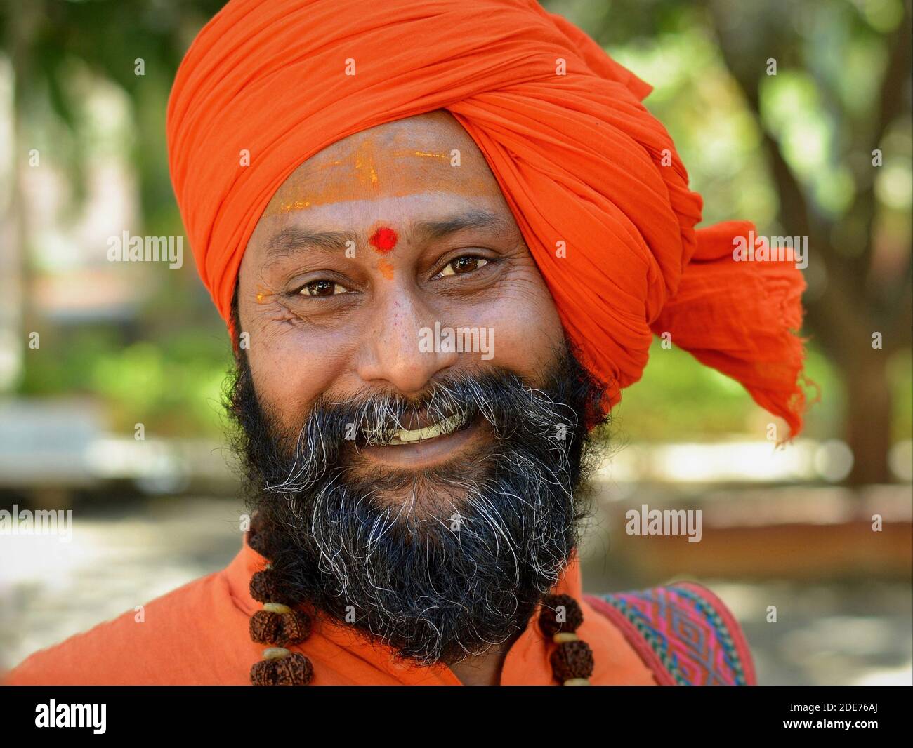 Positiv optimistischer glücklicher junger indischer Heiliger (Sadhu, baba, Mönch) mit orangefarbenem Turban und Rudraksha-Perlen um den Hals lächelt für die Kamera. Stockfoto