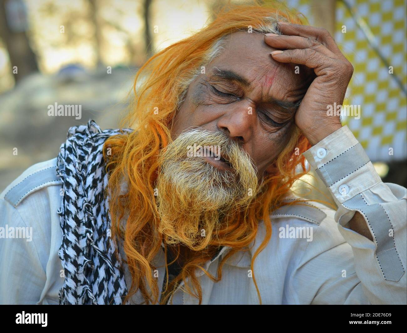 Der alte indische Hindu-Mann mit Henna-gefärbten langen Haaren und langem Bart schläft im Freien bei hellem Tageslicht und legt seine Stirn auf seine linke Hand. Stockfoto