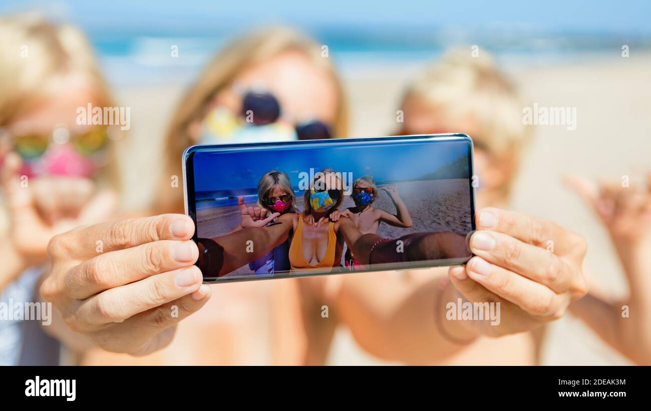 Lustige Kinder mit Mutter machen Selfie-Foto mit dem Smartphone am tropischen Strand. Wearring Gesichtsmaske an öffentlichen Plätzen durch Coronavirus COVID 19 Stockfoto