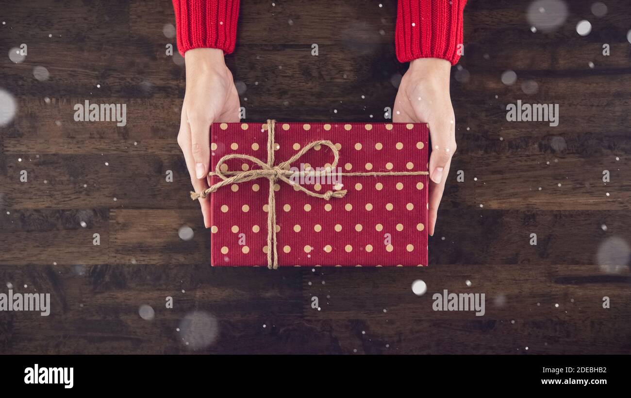 Frau Hände geben christsmas Geschenkbox mit roten gepunkteten verpackt Papier Draufsicht auf Holz Tisch Hintergrund mit Schnee fallen Stockfoto