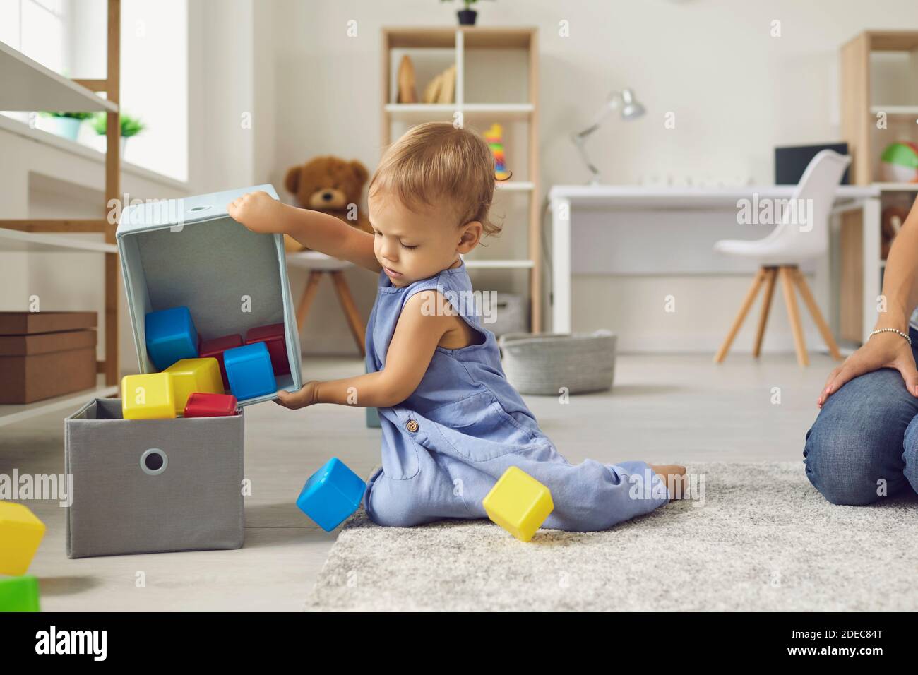 Nettes kleines 2-jähriges Kind, das Spielzeug wieder an ihren Platz setzt und Mama hilft, aufzuräumen Stockfoto