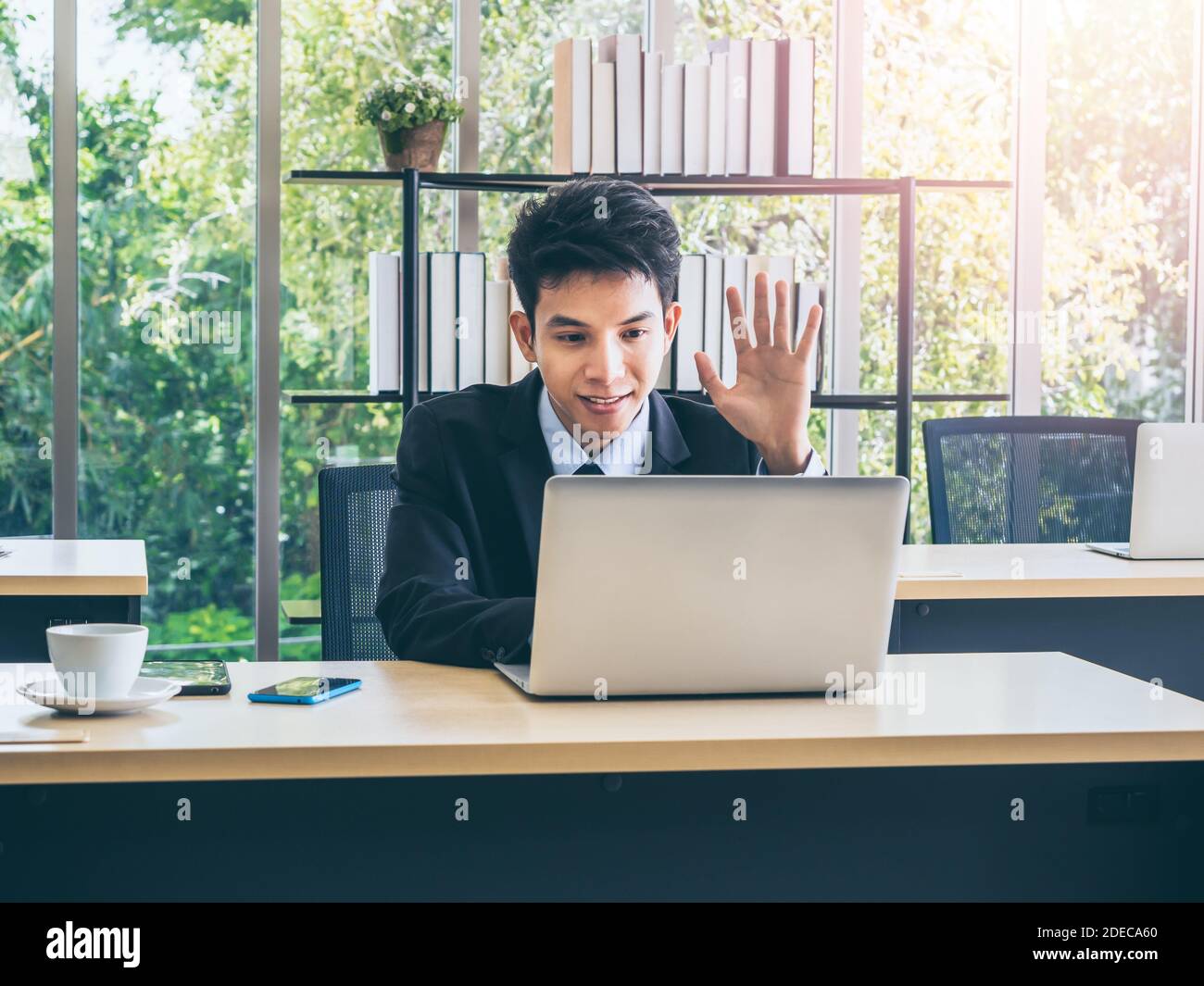 Junge lächelnde asiatische Geschäftsmann in Anzug Gruß jemand, winken zur Kamera auf Laptop-Computer haben Video-Gespräch online während der Arbeit auf Schreibtisch in der Stockfoto