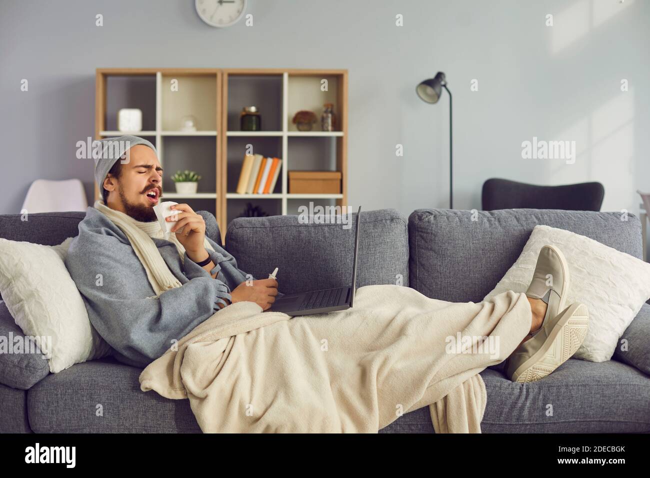 Mann mit laufender Nase und Fieber liegt mit einem Laptop auf der Couch, während er zu Hause isoliert ist. Stockfoto