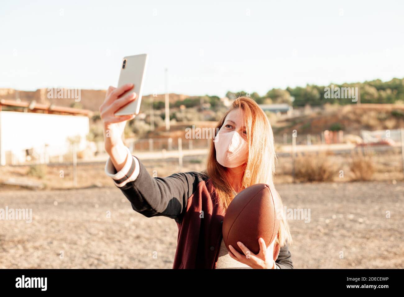 Frau trägt eine schützende Gesichtsmaske und mit ihrem Smartphone, während sie einen american Football Ball hält. Neues normales Konzept. Stockfoto