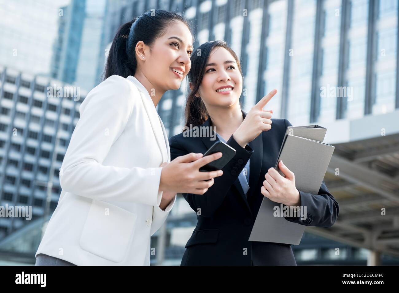 Zwei asiatische weibliche Profis halten Business-Ordner und Smartphone stehen Außerhalb der Bürogebäude der Stadt, um Kollegen oder Kunden zu treffen Stockfoto