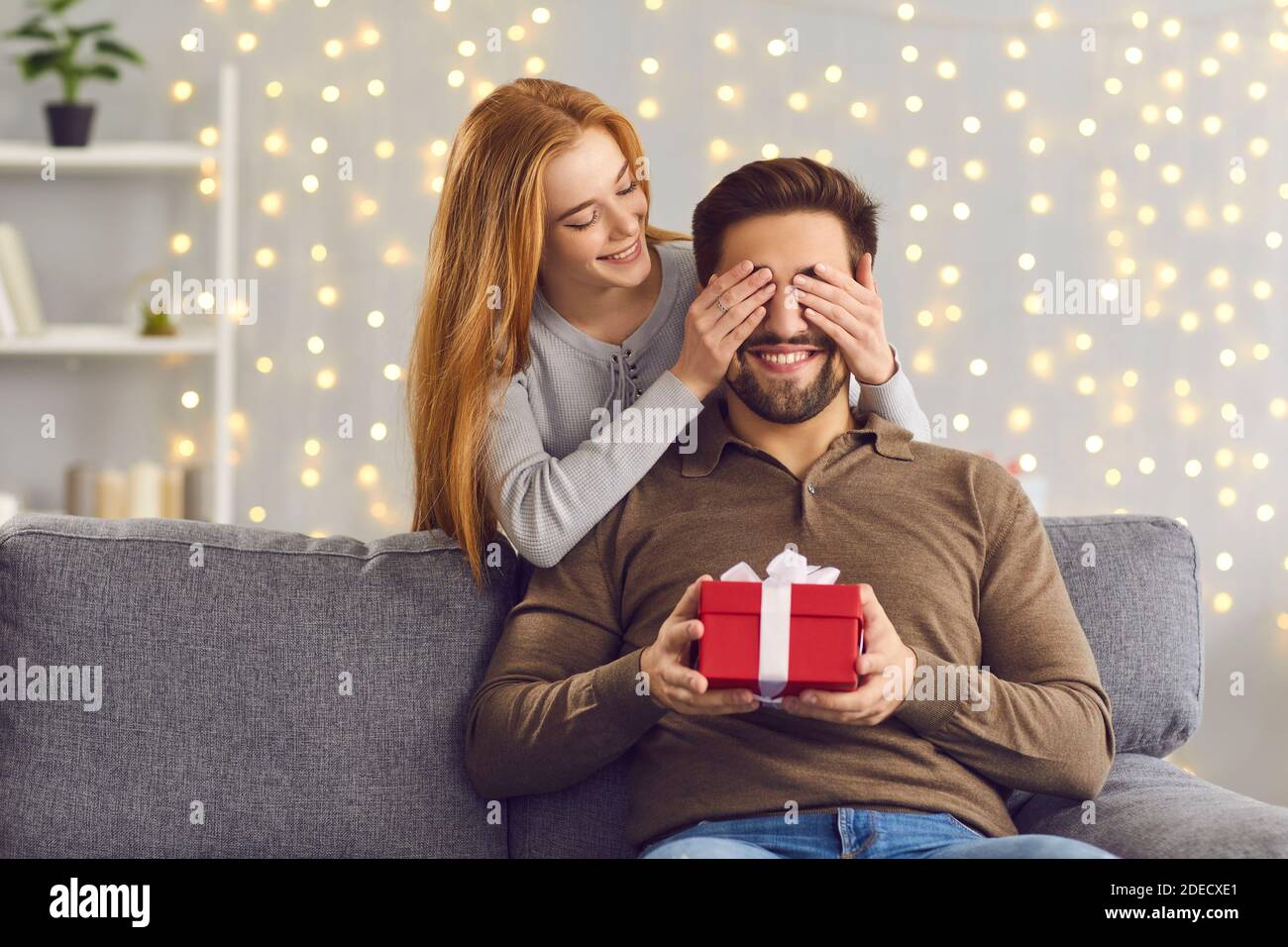 Lächelnde junge Frau bedeckt die Augen ihres Freundes und gibt ihm Ein Überraschungsgeschenk Stockfoto