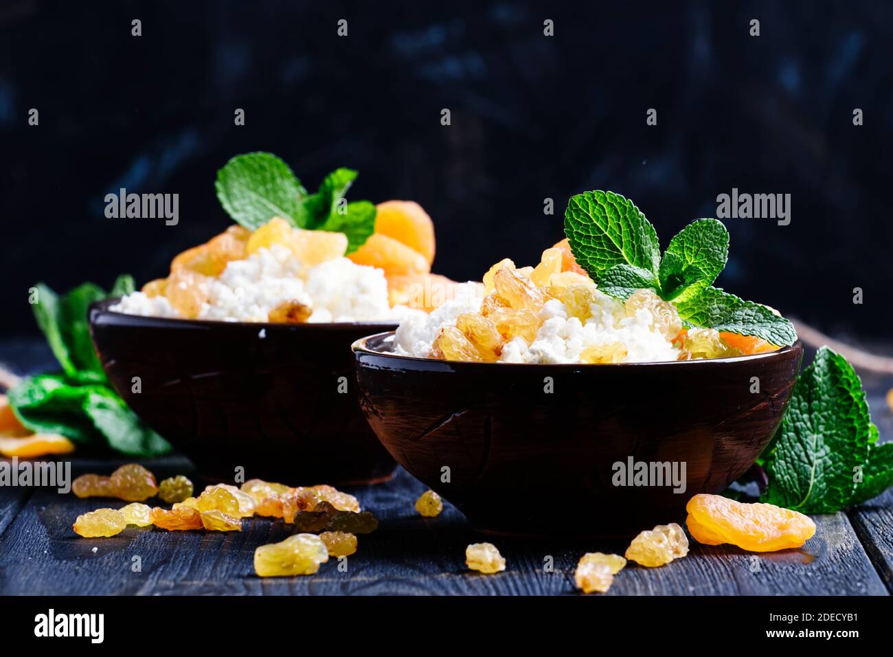 Hausgemachter Hüttenkäse mit getrockneten Aprikosen, Rosinen und Minze in Tonschüsseln, gesundes Frühstück, dunkler Hintergrund, selektiver Fokus Stockfoto