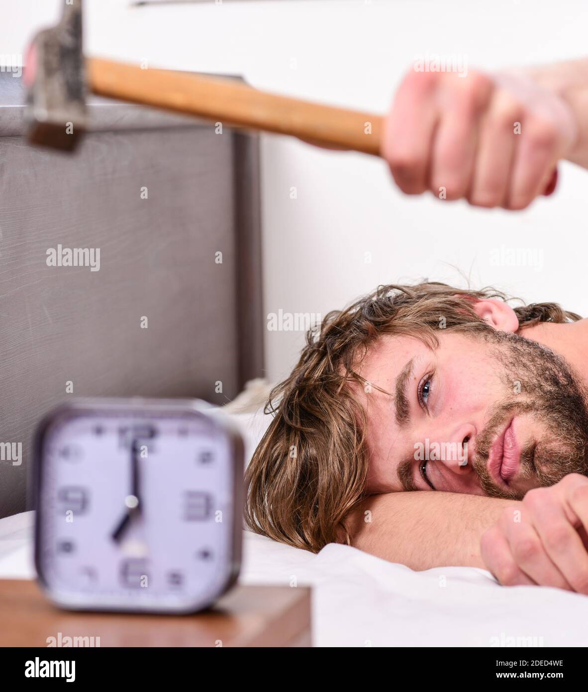 Guy Klopfen mit einem Hammer Wecker klingeln. Pause Disziplin Regime. Ärgerlich Klang. Man bärtige genervt verschlafenes Gesicht legen Kissen in der Nähe von Wecker. Auf zu klingeln. Lästige klingeln Wecker. Stockfoto