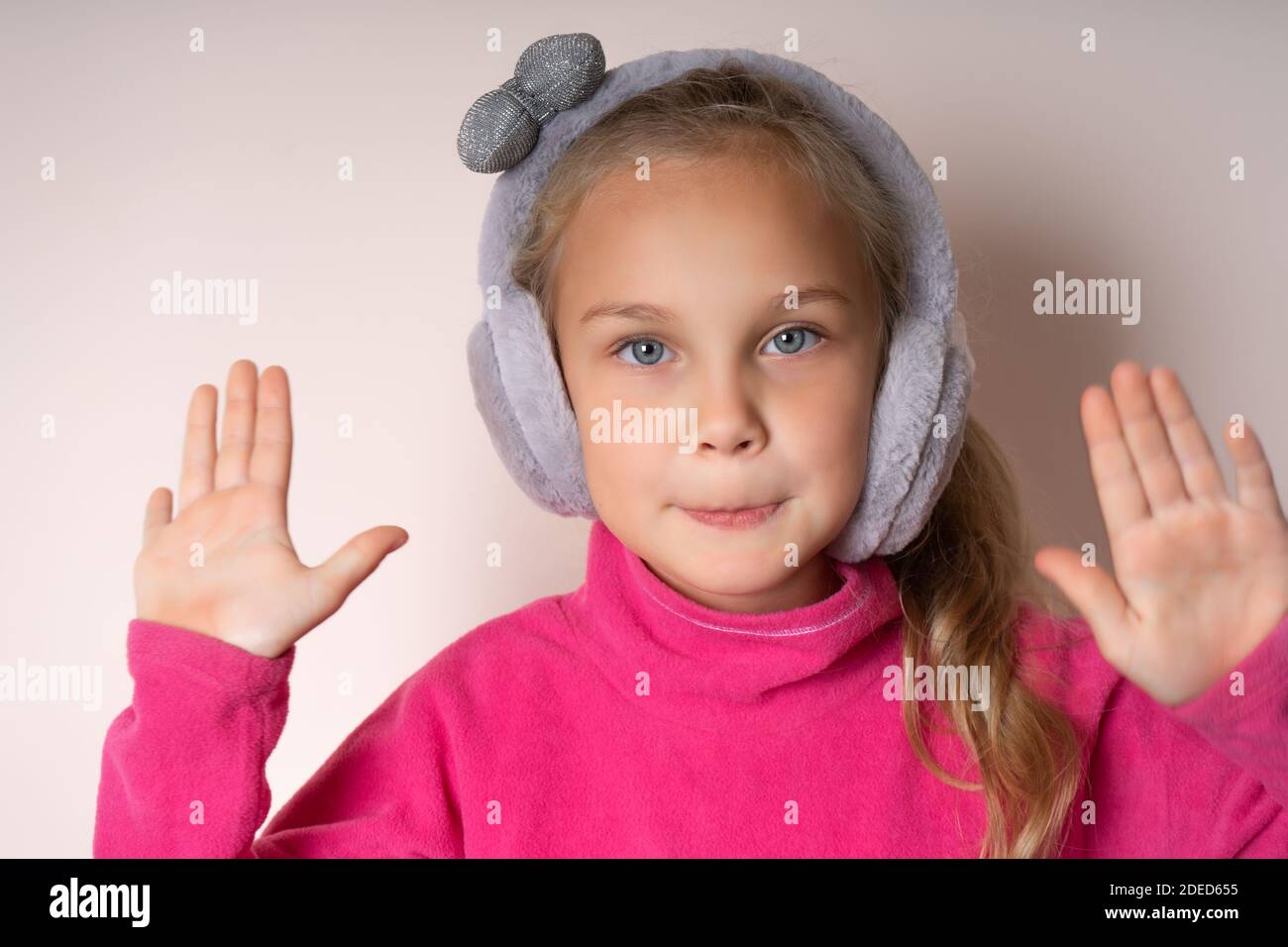 Kleine niedliche Mädchen hob zwei Handflächen nach oben und schaut auf die Kamera, sie ist in warmen Pelz Kopfhörer auf einem hellen Hintergrund Stockfoto