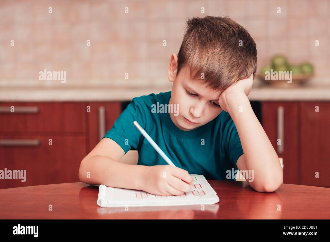 7 yeas alter Junge sitzt am Tisch in der Küche und schreibt Notizen auf Blätter, schwierige Hausaufgaben, Kind konzentriert sich auf Beispiele Stockfoto