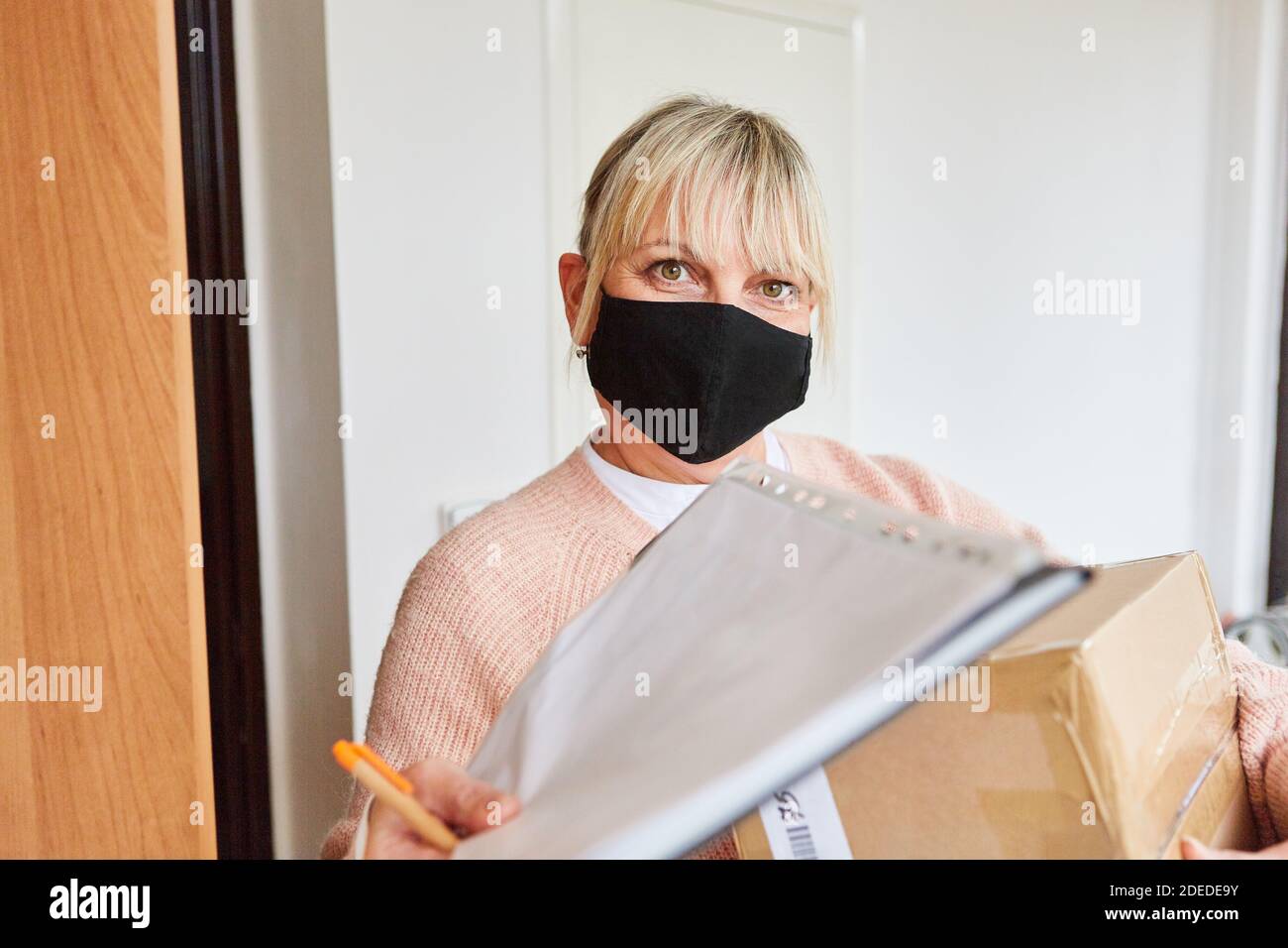 Frau mit Gesichtsmaske vor der Tür von Ihre Wohnung bestätigt die Annahme des Pakets für den Lieferservice Stockfoto
