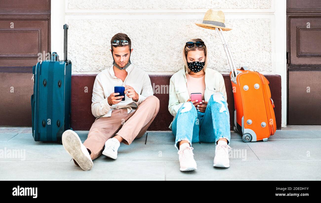 Junges Paar mit Gesichtsmaske mit mobilen Smartphones - Junge Leute, die Online-Reservierungen überprüfen, sitzen auf dem Boden vor dem Zug Station Stockfoto