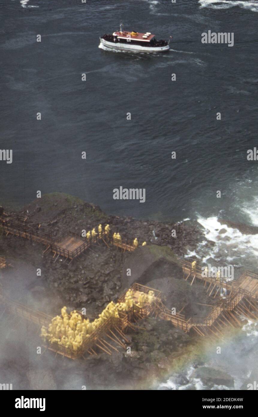 1970s Photo (1973) - bei Cave of the Winds am Fuß der Klippen auf Bootsinsel blicken Touristen in gelber Regenkleidung auf die amerikanischen Stürze aus dem Fluss. Ein Ausflugsboot der Maid-of-the-Mist liegt zwischen der amerikanischen und kanadischen Küste. Stockfoto