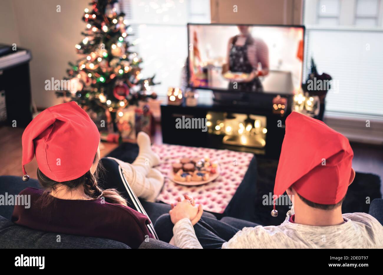 Ein Paar schaut zu Weihnachten fern. Glücklicher Familienurlaub zu Hause. Mann und Frau auf der Couch entspannen mit Baum, Dekorationen, Lichter, Kerzen und Fernsehen. Stockfoto