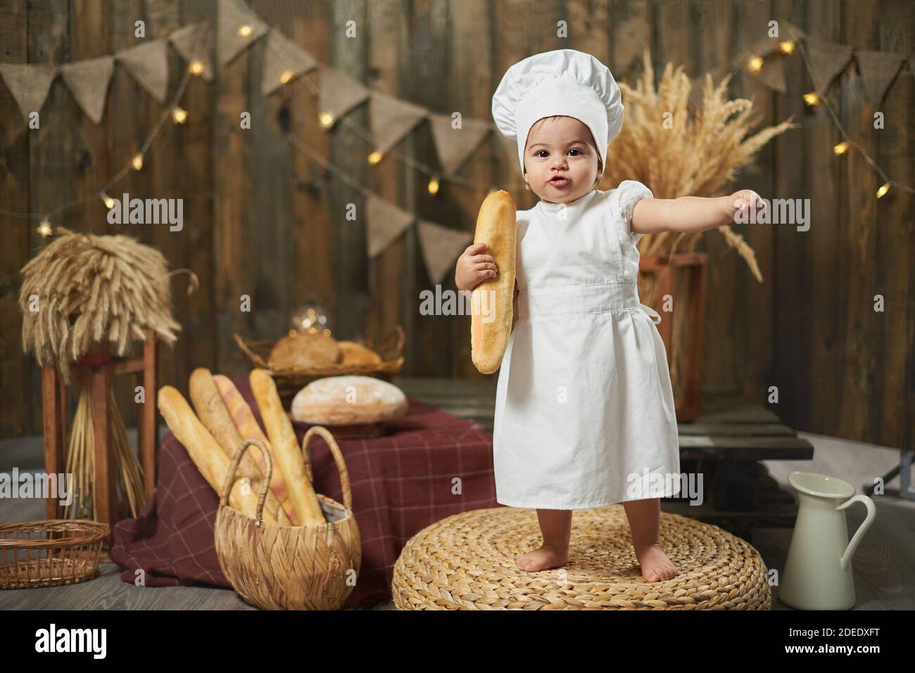Niedliches kleines Baby trägt ein Bäckerkostüm mit einem französischen Baguette In einem rustikalen Interieur Stockfoto