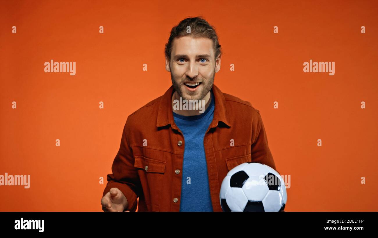 Positiver Fußballfan, der die Kamera anschaut, während er den Fußball hält Auf orange Stockfoto