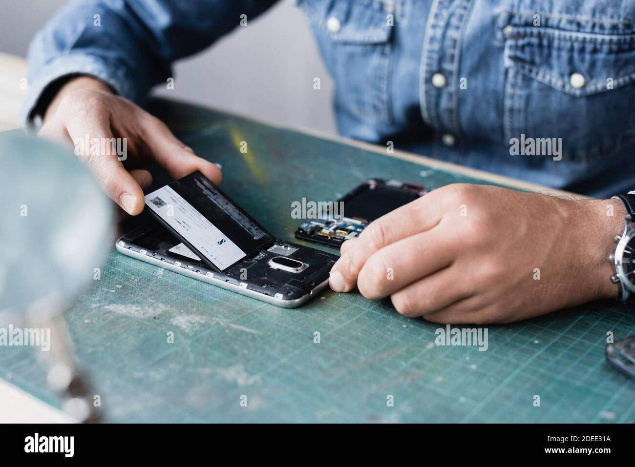 Beschnittene Ansicht des Reparaturwerkers, der den Akku in das Mobiltelefon eingelegt hat Tabelle mit verschwommener Lupe im Vordergrund Stockfoto