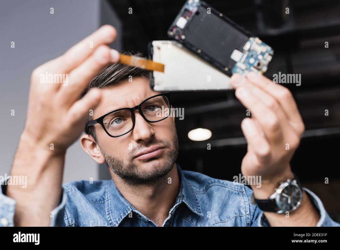Unzufriedener Reparaturman in Brillen Blick auf zerlegte Teile gebrochen Mobiltelefon im unscharfen Vordergrund Stockfoto