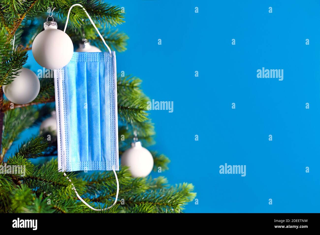 Medizinische Gesichtsmaske, die vom Weihnachtsbaum auf blauem Hintergrund mit leerem Kopierraum hängt. Konzept für Weihnachtsfeier in Zeiten des Corona Virus Stockfoto