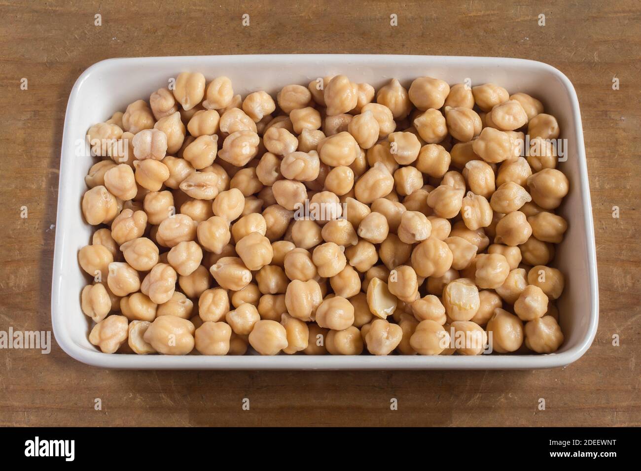 Eine weiße Keramikschale voller roher weißer Kichererbsen mit einem gelblichen Ton, nachdem sie auf einem Holztisch eingeweicht wurde. Organische Texturen als Hintergrund. Stockfoto