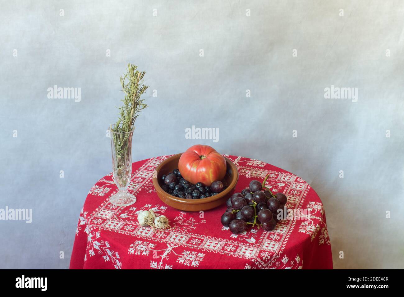 Stillleben einer Steingutschüssel voller Früchte, Gemüse, Nüsse auf einem Tisch mit einer roten Tischdecke mit Weihnachtsschmuck und Rosmarin bedeckt. Stockfoto