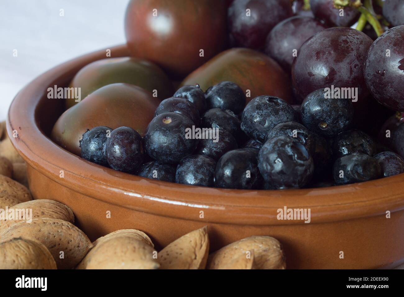 Nahaufnahme von einigen Blaubeeren mit einem Bund schwarzer Trauben, Tomaten und Mandeln in einer Tonschüssel auf einem Küchentisch in einem Haus. Stockfoto
