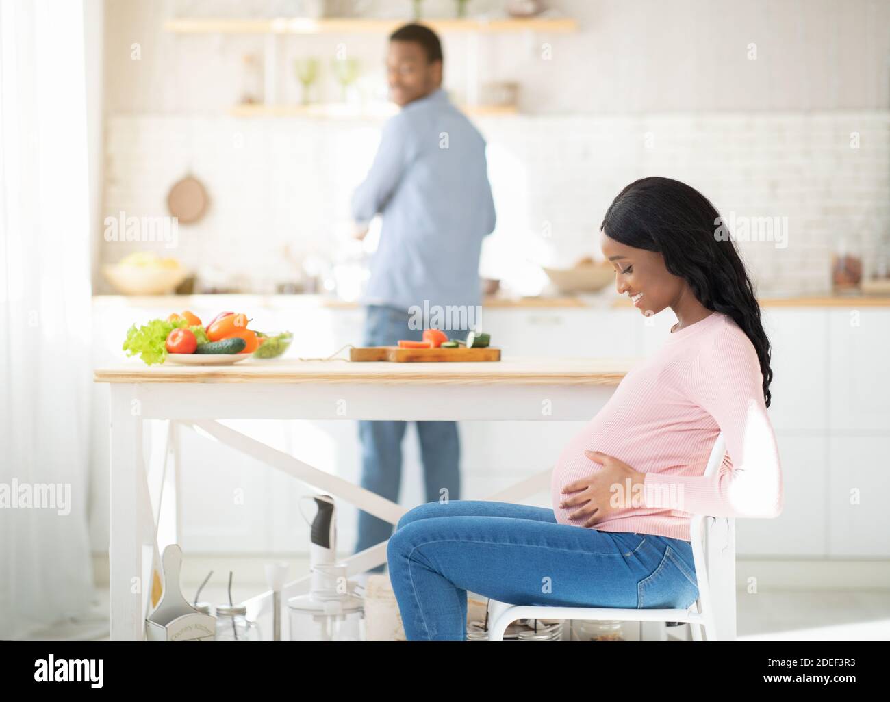 Gesunde Ernährung während der Schwangerschaft. Schöne werdende Dame berühren ihren Bauch, während Mann Kochen Mittagessen in der Küche Stockfoto