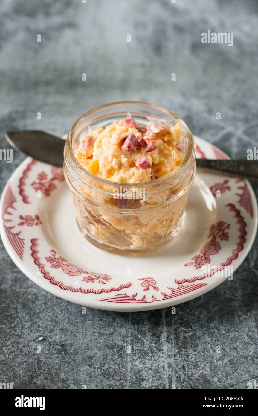 Hausgemachter Pimento-Käse in einem Glasbehälter, auf einem Teller mit einem Messer vor grauem Hintergrund. Nahaufnahme. Stockfoto