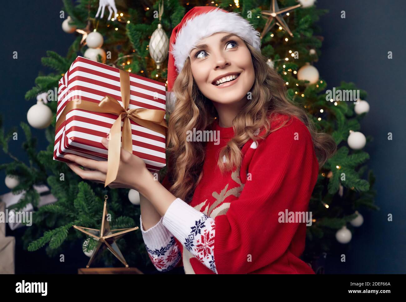 Portrait von wunderschönen blonden Frau in roten Pullover und Santa Hut hält ein Geschenk in der weihnachtlich dekorierten Innenraum. Stockfoto