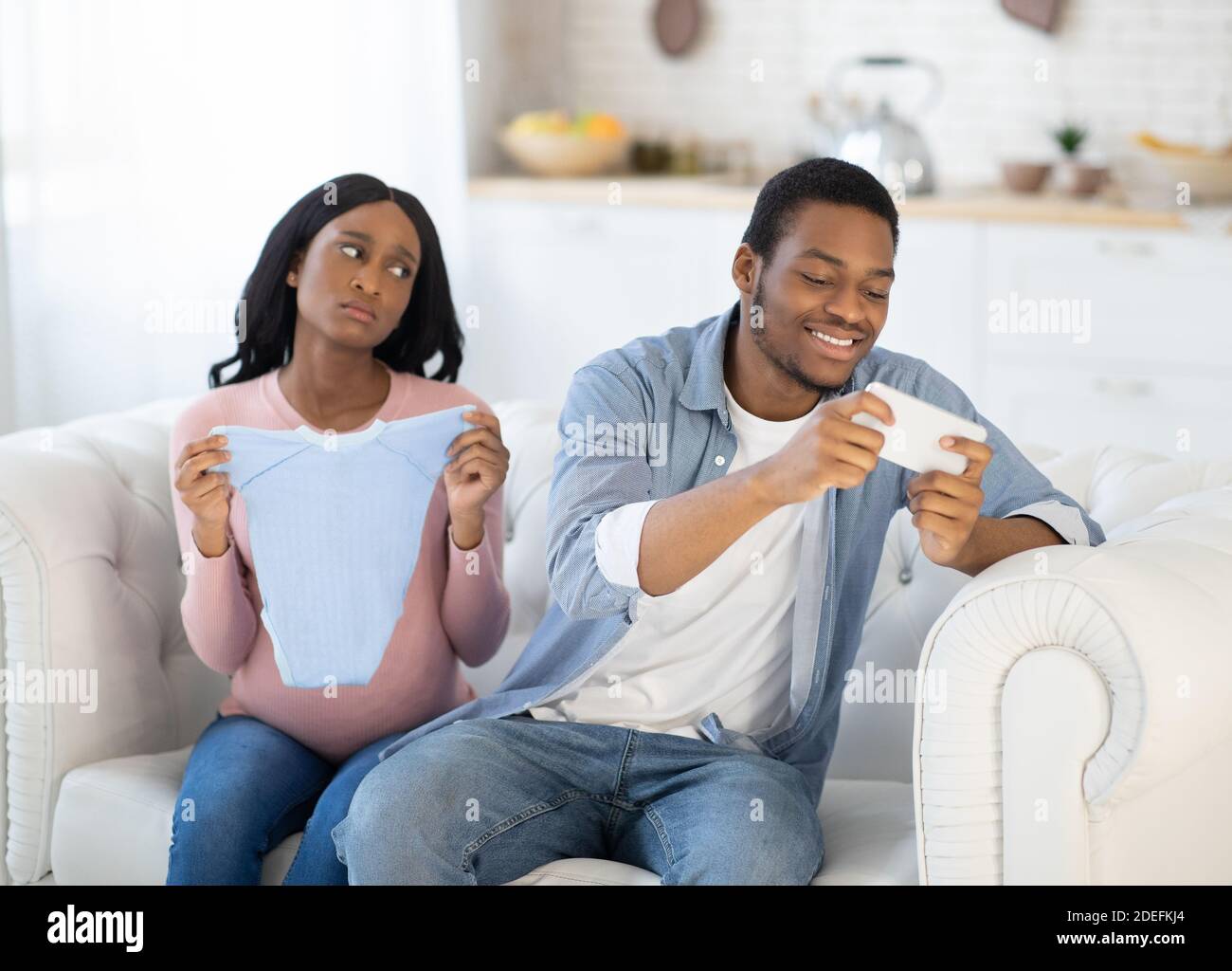 Gadget sucht Konzept. Egoistischer schwarzer Mann, der Spiele auf dem Handy spielt und seine erwartungsvolle Frau zu Hause vernachlässigt Stockfoto