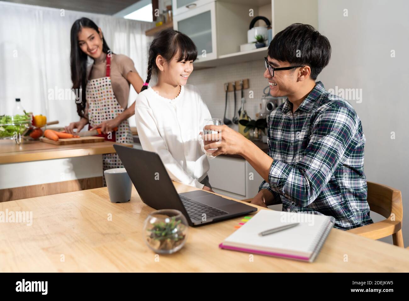 Asiatische kleine Mädchen, das Wasser zu ihrem Vater während der Arbeit mit Computer-Laptop und Mama Kochen in der Küche. Familienarbeit zu Hause während Quarantäne von CO Stockfoto