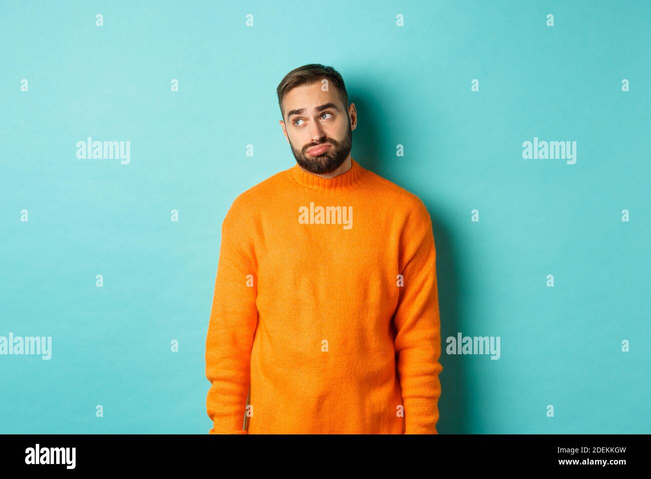 Trauriger und düsterer Mann schmollend, langweilen in der oberen linken Ecke, in orangefarbenem Pullover vor hellblauem Hintergrund stehend Stockfoto