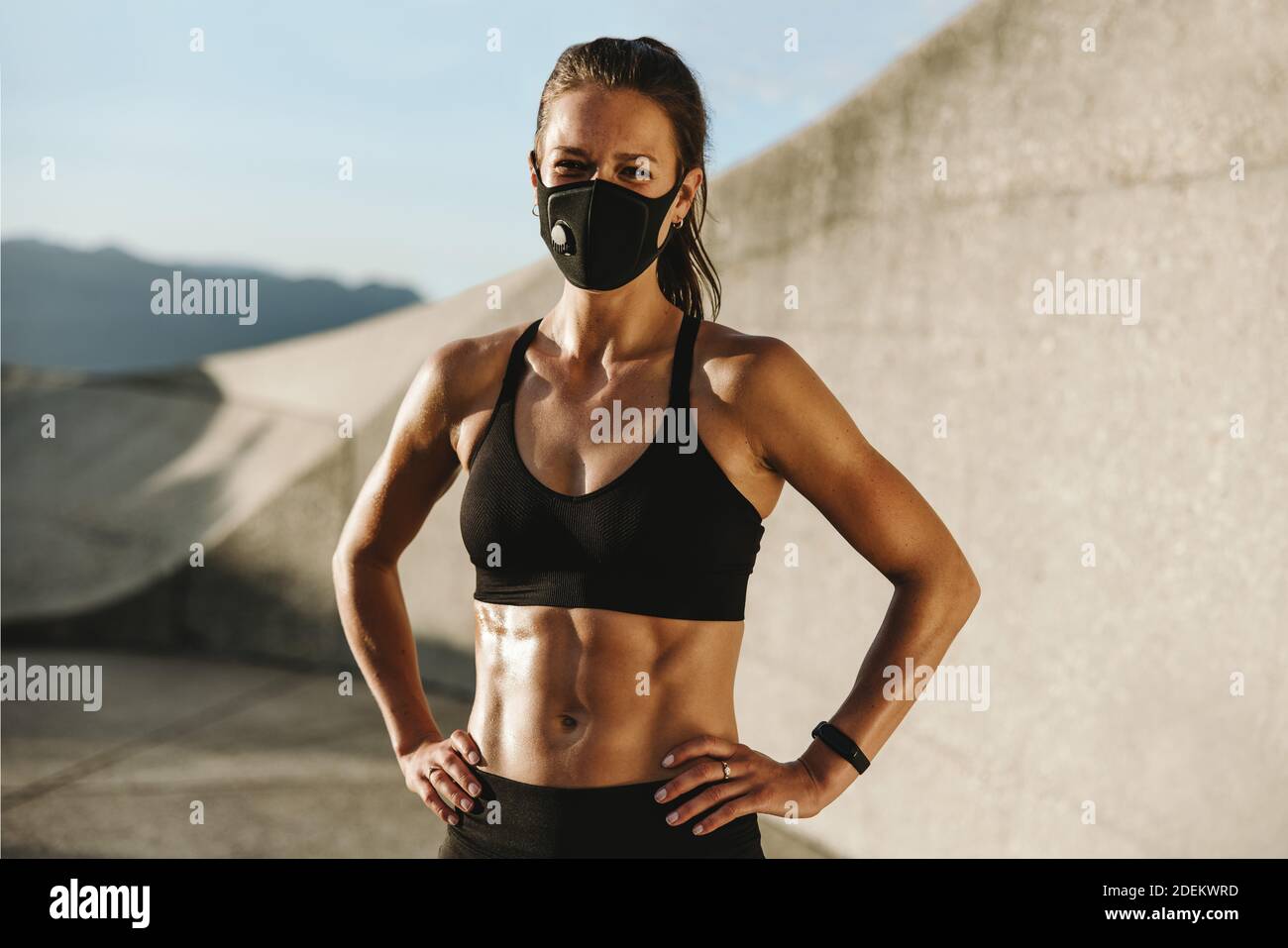 Fit Frau trägt schützende Gesichtsmaske Blick auf die Kamera. Hündin in Sportswear, die morgens mit den Händen auf den Hüften im Freien steht. Stockfoto