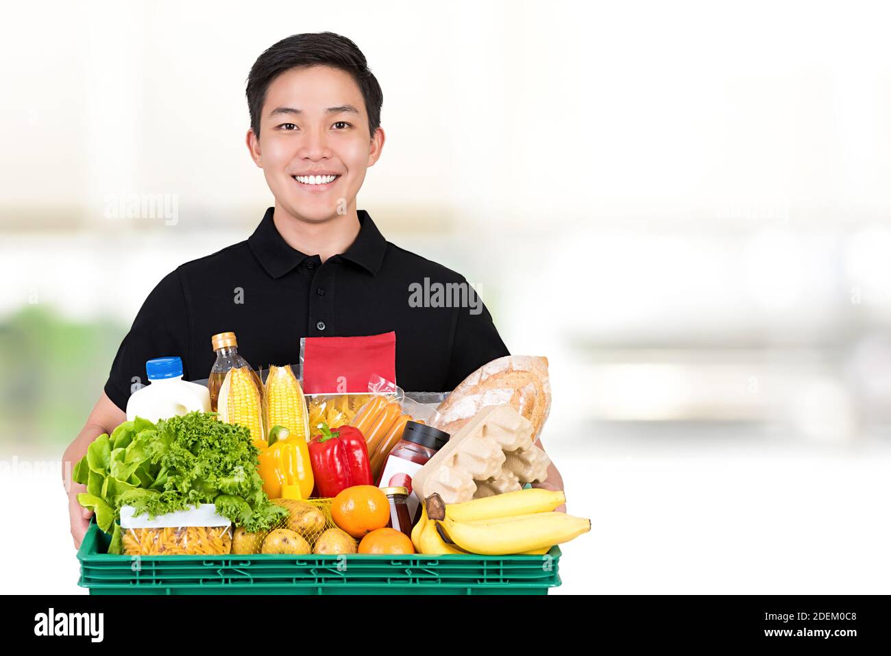 Lächelnder Lebensmittelladen asiatischer Liefermann trägt ein schwarzes Poloshirt Halten des Lebensmittelkorbs auf weißem Hintergrund mit Kopierplatz Stockfoto