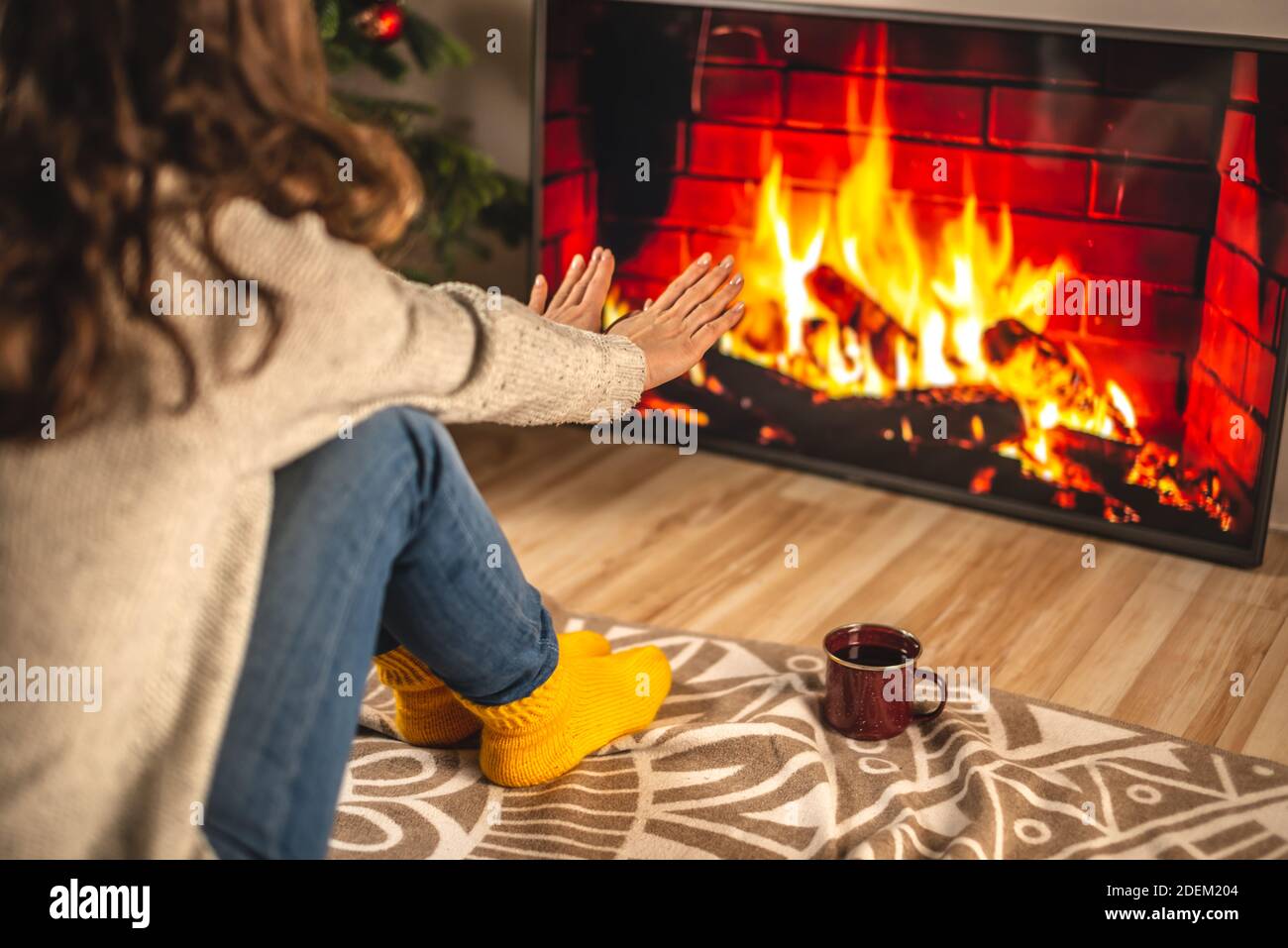 Frau in einem Pullover und gelben warmen Socken sitzt vor dem Kamin, der auf dem TV-Bildschirm dargestellt ist und wärmt ihre Hände. CR-Konzept Stockfoto