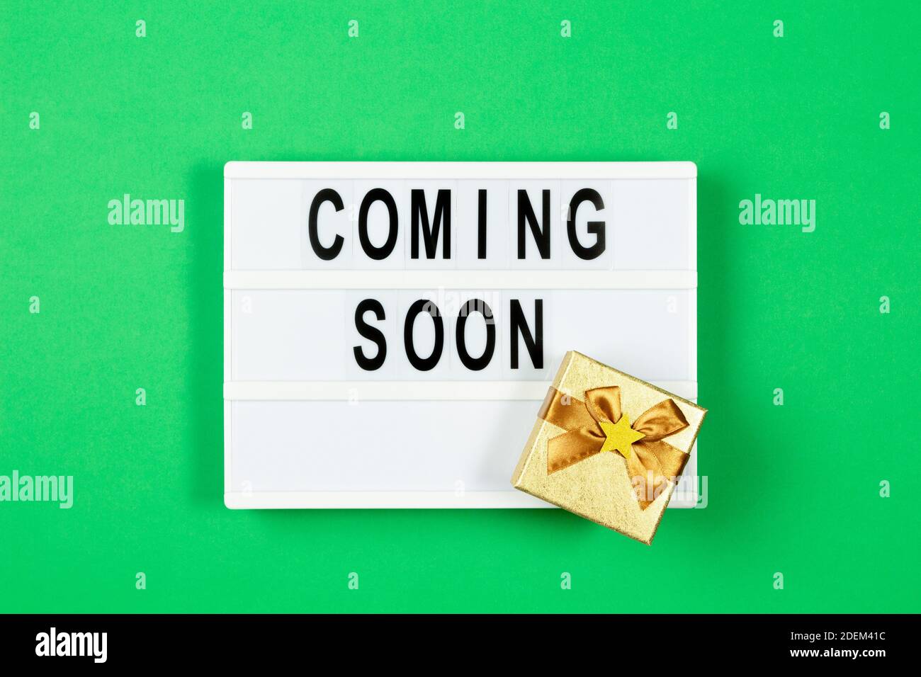 Weißer Leuchtkasten mit Text IN KÜRZE und kleine Gold Geschenk-Box auf grünem Hintergrund. Draufsicht Stockfoto