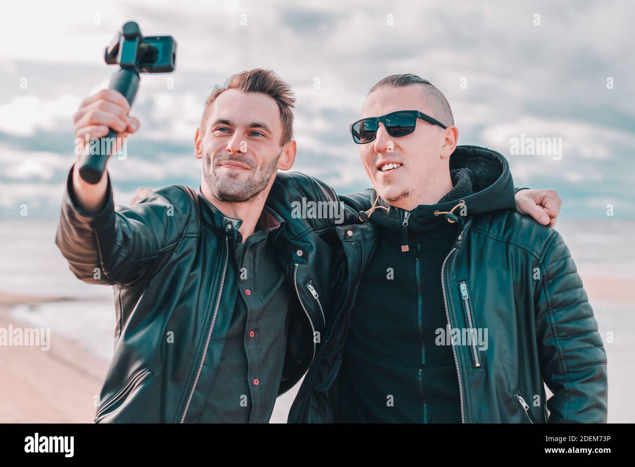 Zwei gutaussehende lächelnde Freunde machen Selfie mit Action-Kamera mit Gimbal Stabilisator am Strand. Jugendliche Männer in schwarzen Kleidern Spaß haben, indem Sie Fotos Stockfoto