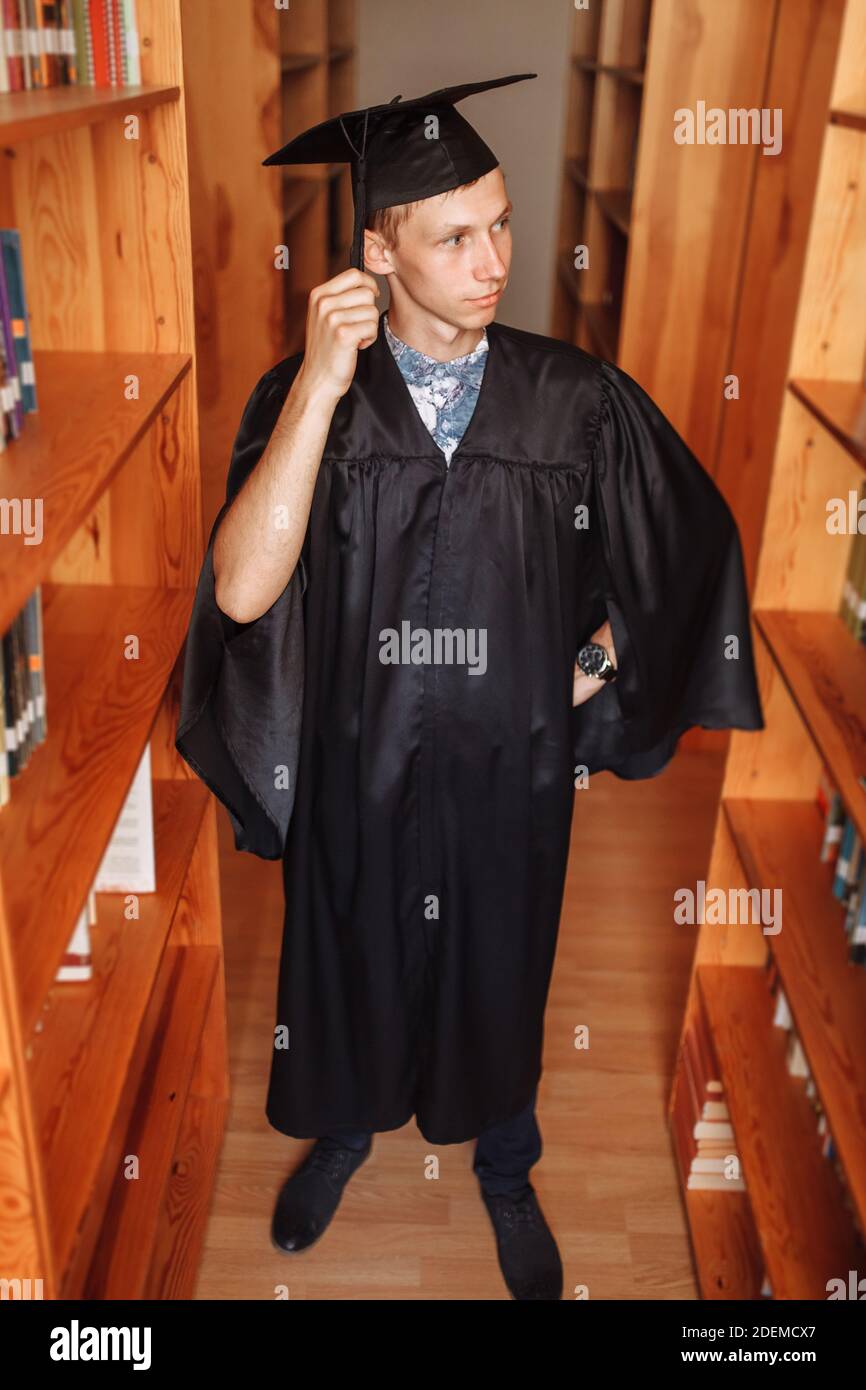 Erfolgreicher Absolvent Kerl, in akademischen Kleidern, in der Bibliothek stehend, Bücher wählend, kann für Werbung verwendet werden, Stockfoto