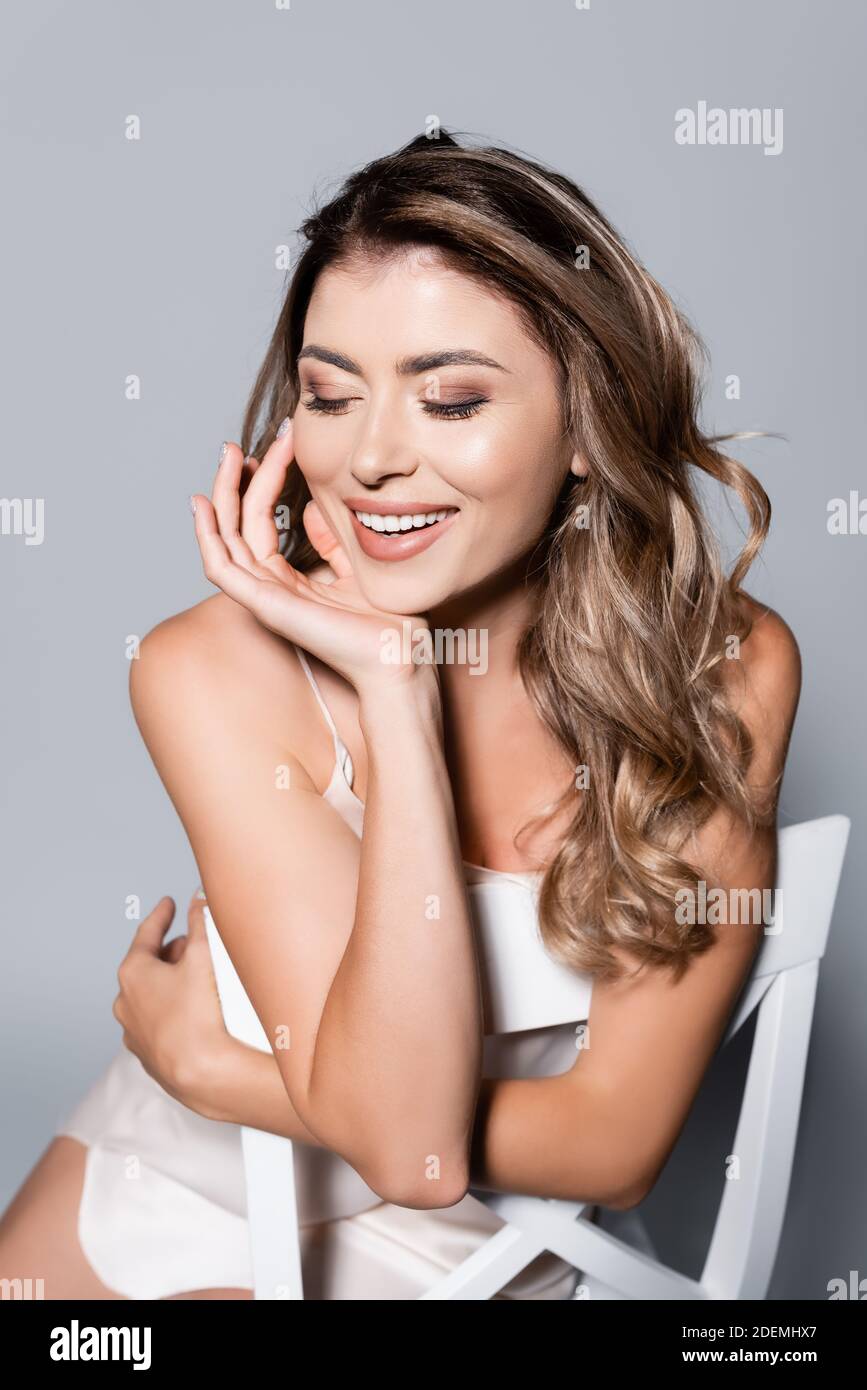 Lächelnd elegante Frau im Kleid posiert auf Stuhl isoliert auf Grau Stockfoto