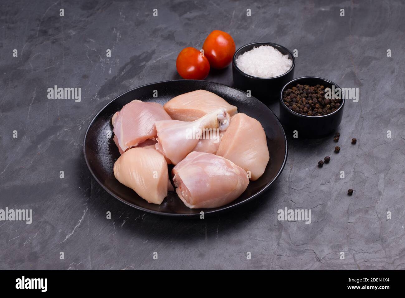 Rohes Huhn zartes Curry ohne Haut auf schwarzem Teller angeordnet geschnitten und garniert mit, Tomaten, schwarzem Pfeffer und Salz auf Stein texturierten Hintergrund Stockfoto