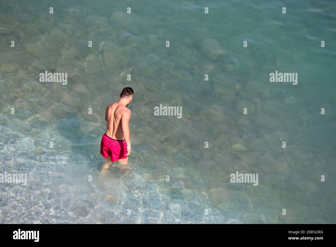 Mann in roten Shorts geht in einem Meer schwimmen. Luftaufnahme zum Kiesstrand mit transparentem Wasser, Hintergrund für Urlaub und Reisen Stockfoto