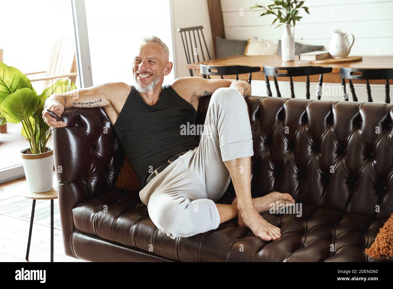 Schöner glücklicher Mann mittleren Alters in legerer Kleidung mit Smartphone in der Hand entspannen auf bequemen Sofa in seiner modernen Wohnung, Blick beiseite und Stockfoto