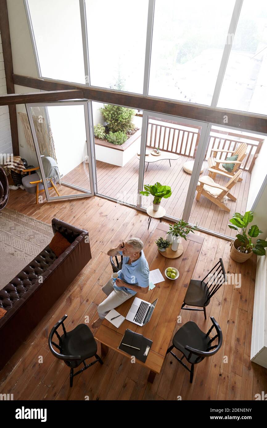 Vertikale Aufnahme eines kaukasischen Geschäftsmannes mittleren Alters, der mit dem Mobiltelefon spricht, während er aus der Ferne in seiner modernen Wohnung arbeitet, Draufsicht Stockfoto