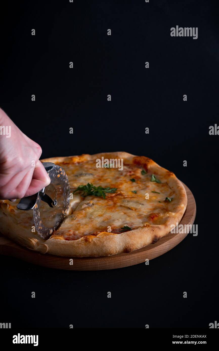 Eine Nahaufnahme einer Person, die einen frisch gebackenen Käsekitsch schneidet Pizza auf einem Holztablett auf schwarzem Hintergrund Stockfoto