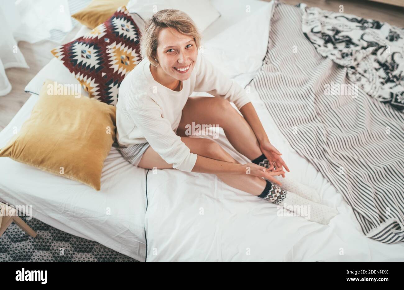Junge blonde Frau in Pyjama gekleidet sitzen im gemütlichen Bett mit Kissen und aufrichtig lächelnd und Blick auf die Kamera. Wohnen zu Hause und süß gemütlich Stockfoto