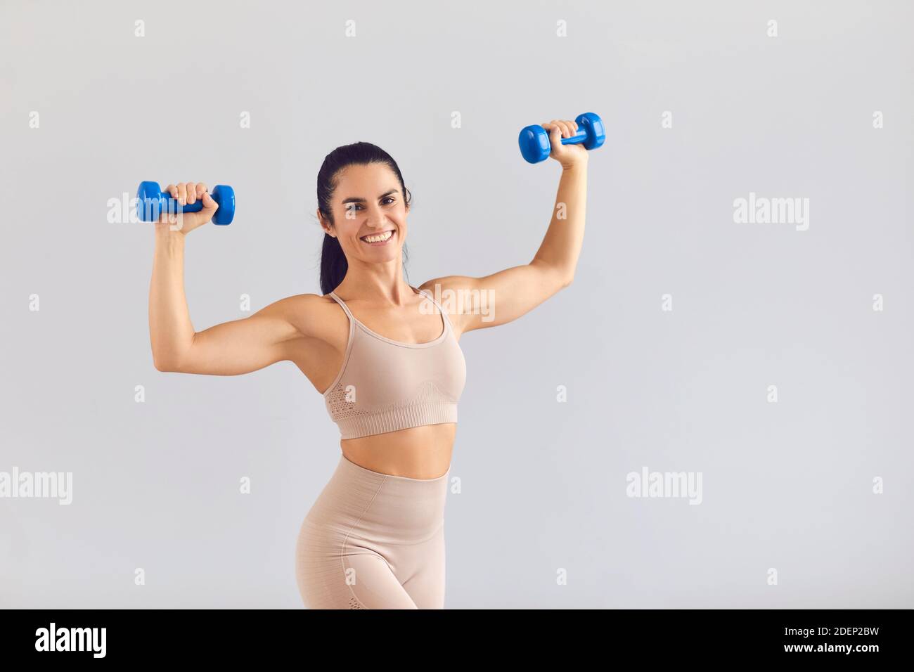 Positive sportliche Frau in beige activewear lächelnd und posiert mit Kurzhanteln im Studio Stockfoto